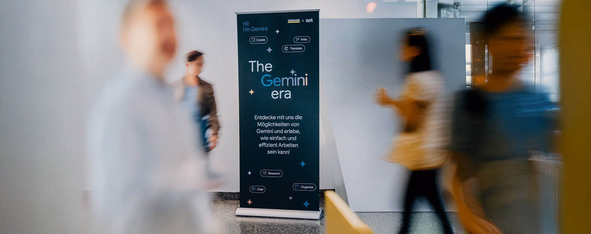Banner in a hallway reading 'The Gemini era' with blurred people walking around it.