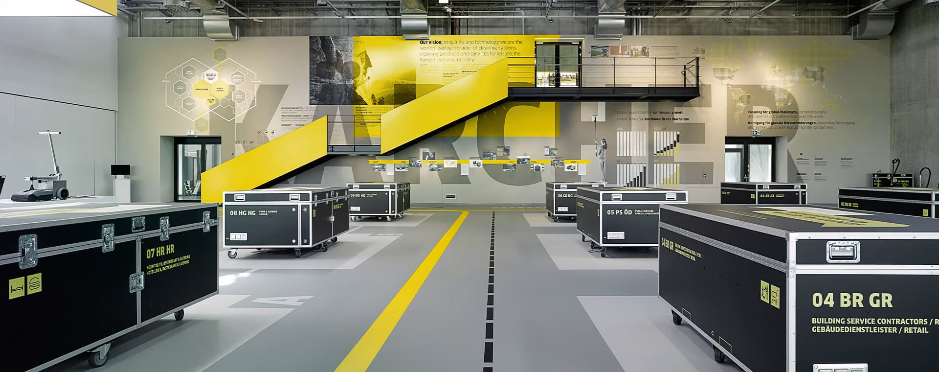 Modern industrial space at Kärcher with black equipment cases on wheels and yellow stairs against informative wall graphics.