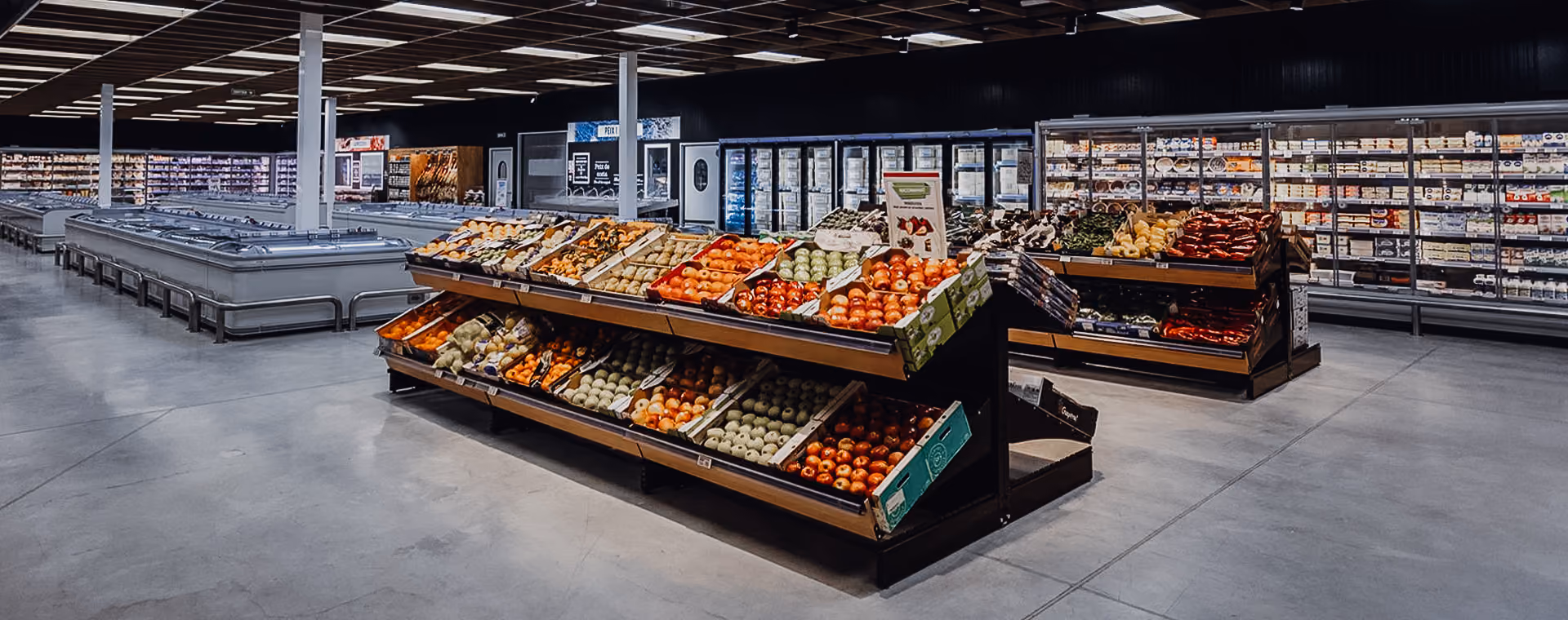 Spacious grocery store produce section with fresh fruits and vegetables on display under bright lights.