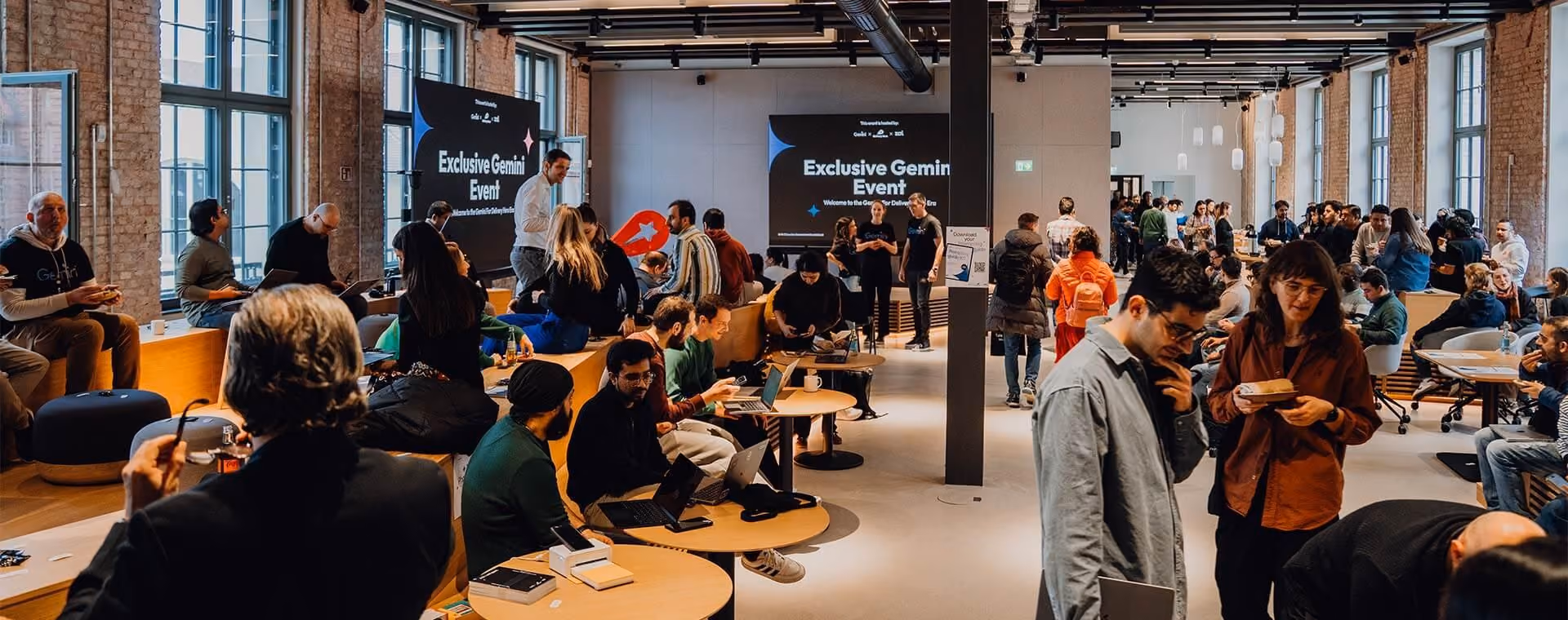 Crowded modern event space with people socializing, working on laptops, and sitting on benches.
