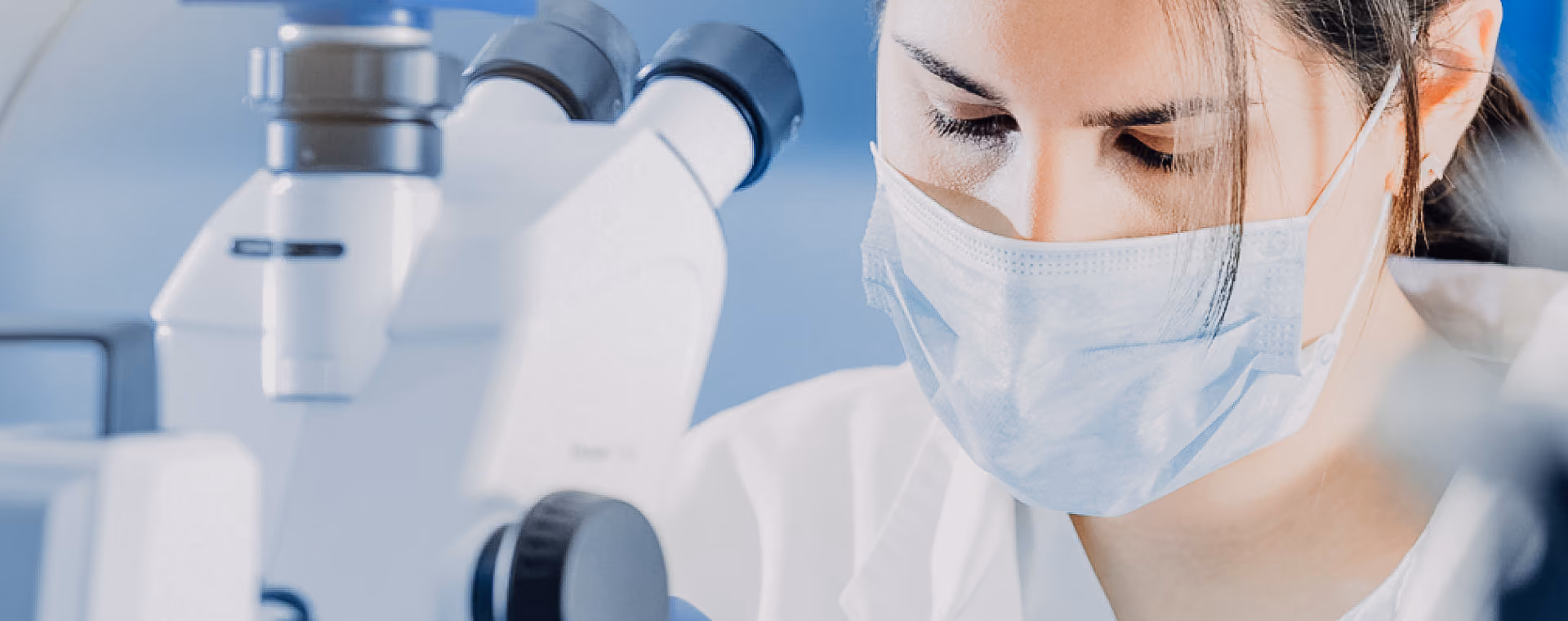 Scientist wearing a mask looking through a microscope in a laboratory setting.