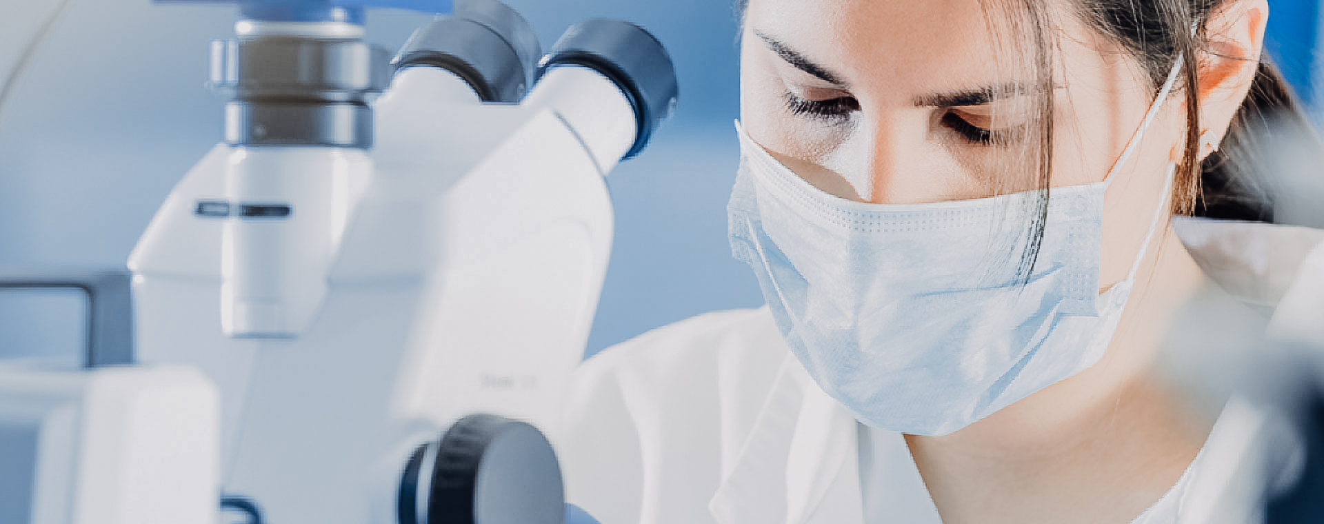 Wissenschaftlerin mit Maske, die in einer Laborumgebung durch ein Mikroskop schaut.