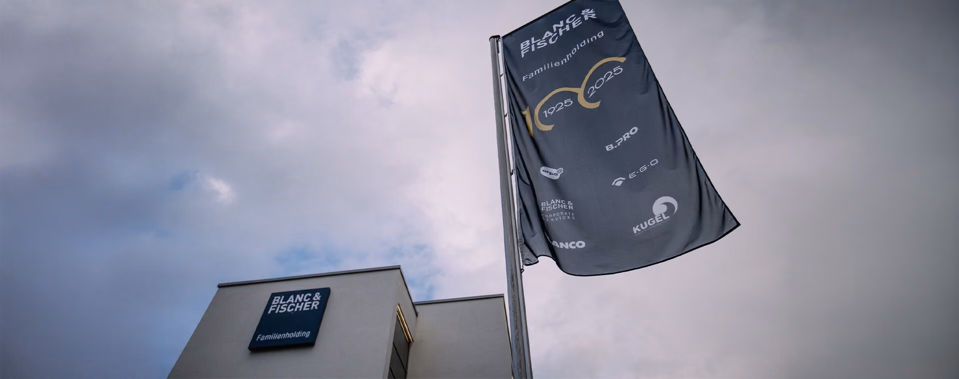 Flag and building facade displaying Blanc & Fischer family holding logos under a cloudy sky.
