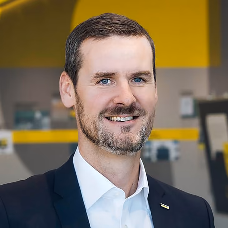 Lächelnder Mann mit kurzen dunklen Haaren und Bart, der eine Anzugjacke und ein weißes Hemd trägt.