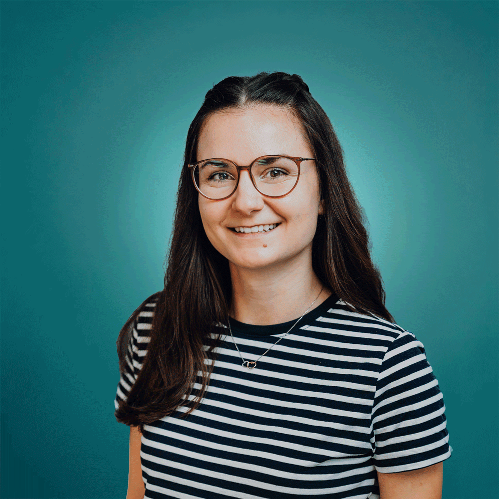 Smiling woman with long dark hair, glasses, and a striped black and white shirt against a teal background.