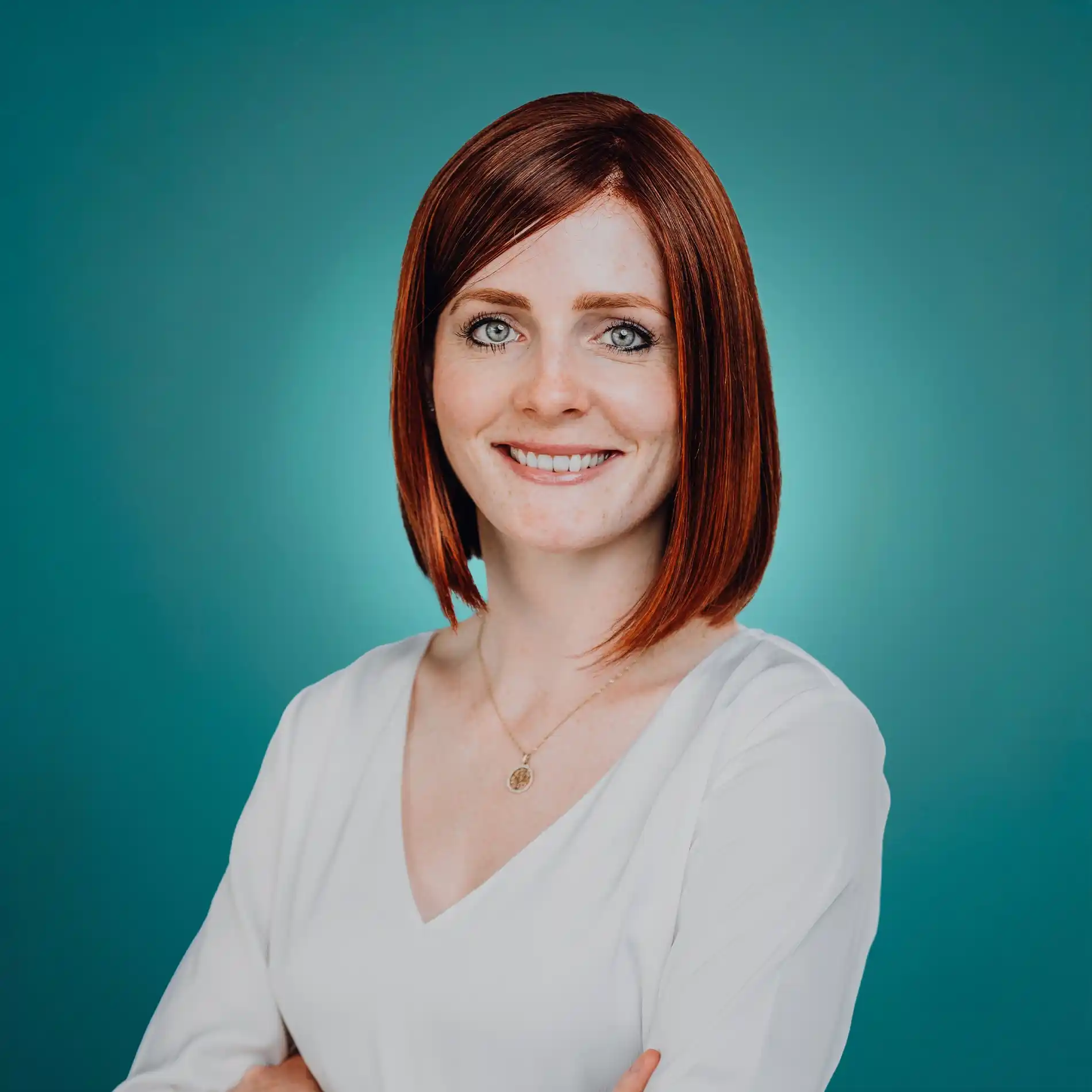 Smiling woman with red bob haircut in white blouse against teal background, arms crossed.