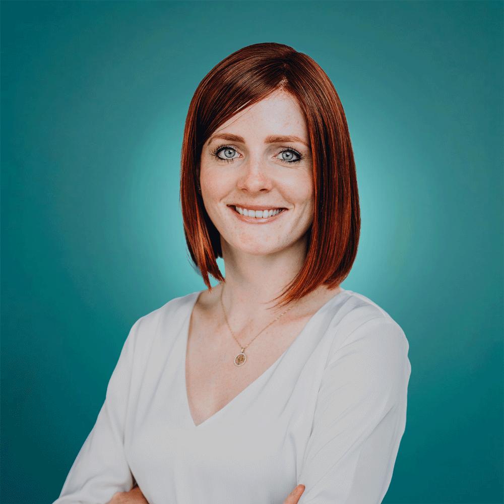 Smiling woman with red bob haircut in white blouse against teal background, arms crossed.