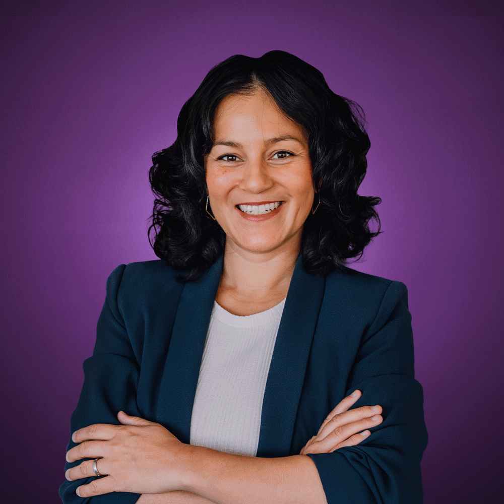 Smiling woman with dark curly hair in a dark blazer and white top crossed arms on purple background.