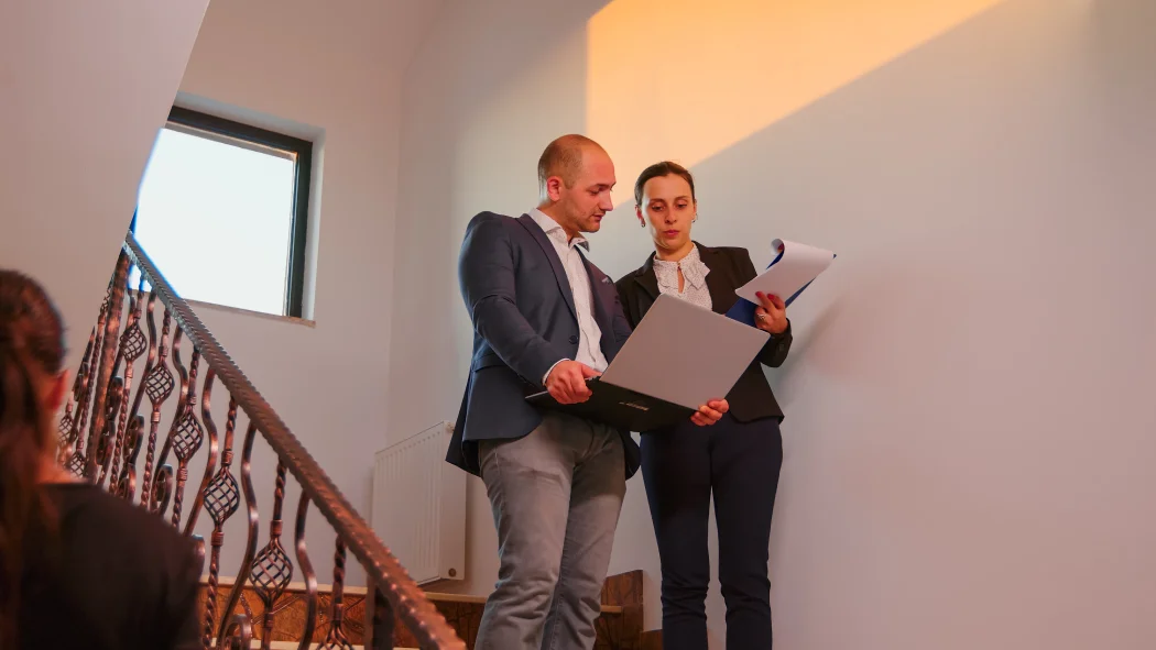 eux professionnels de l'immobilier consultant un ordinateur portable et un dossier dans la cage d'escalier d'un immeuble, illustrant la vérification des garanties légales de la loi Hoguet.