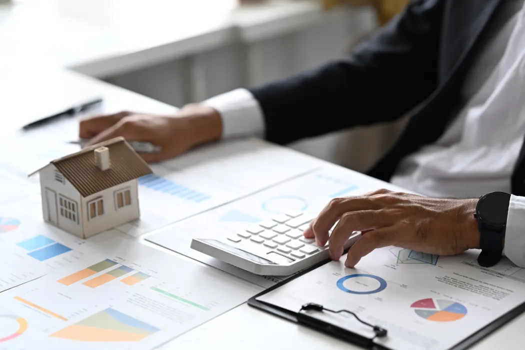 Un investisseur en costume utilisant une calculatrice sur un bureau couvert de graphiques financiers à côté d'une maquette de maison, illustrant le calcul de rentabilité pour un investissement locatif sans apport.