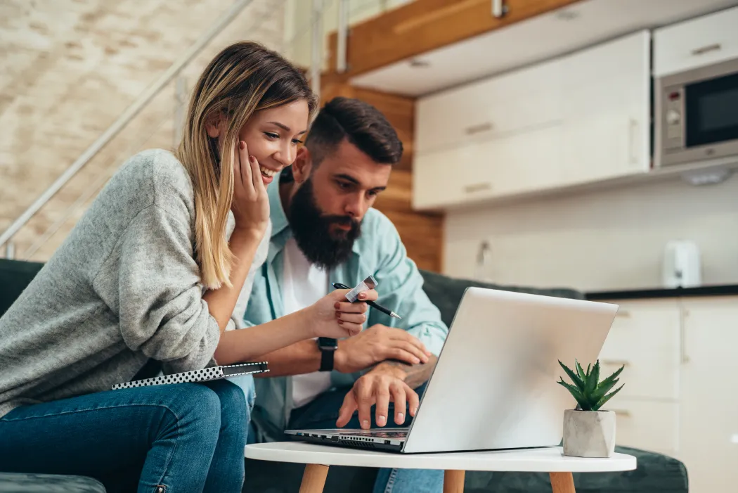 Un jeune couple souriant consultant un ordinateur portable sur un canapé pour suivre sa demande de crédit immobilier et vérifier sa condition suspensive de prêt.