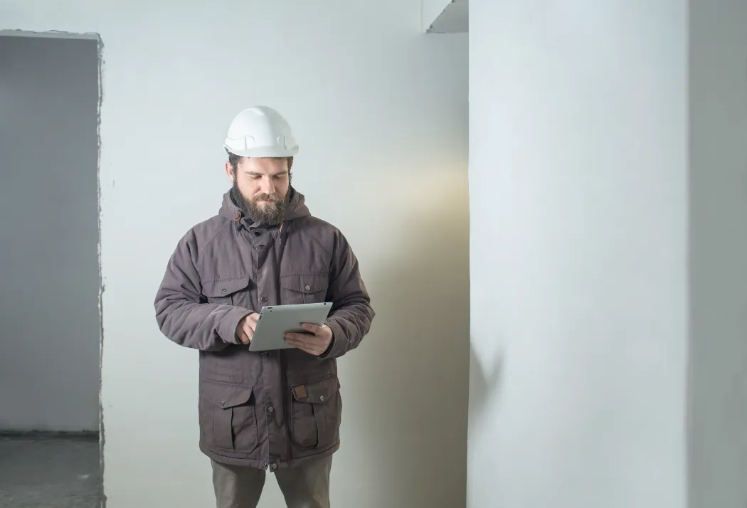 Un expert du bâtiment équipé d'un casque blanc contrôlant la structure d'un logement sur sa tablette numérique, illustrant l'expertise technique recommandée pour détecter ou prouver un vice caché immobilier.
