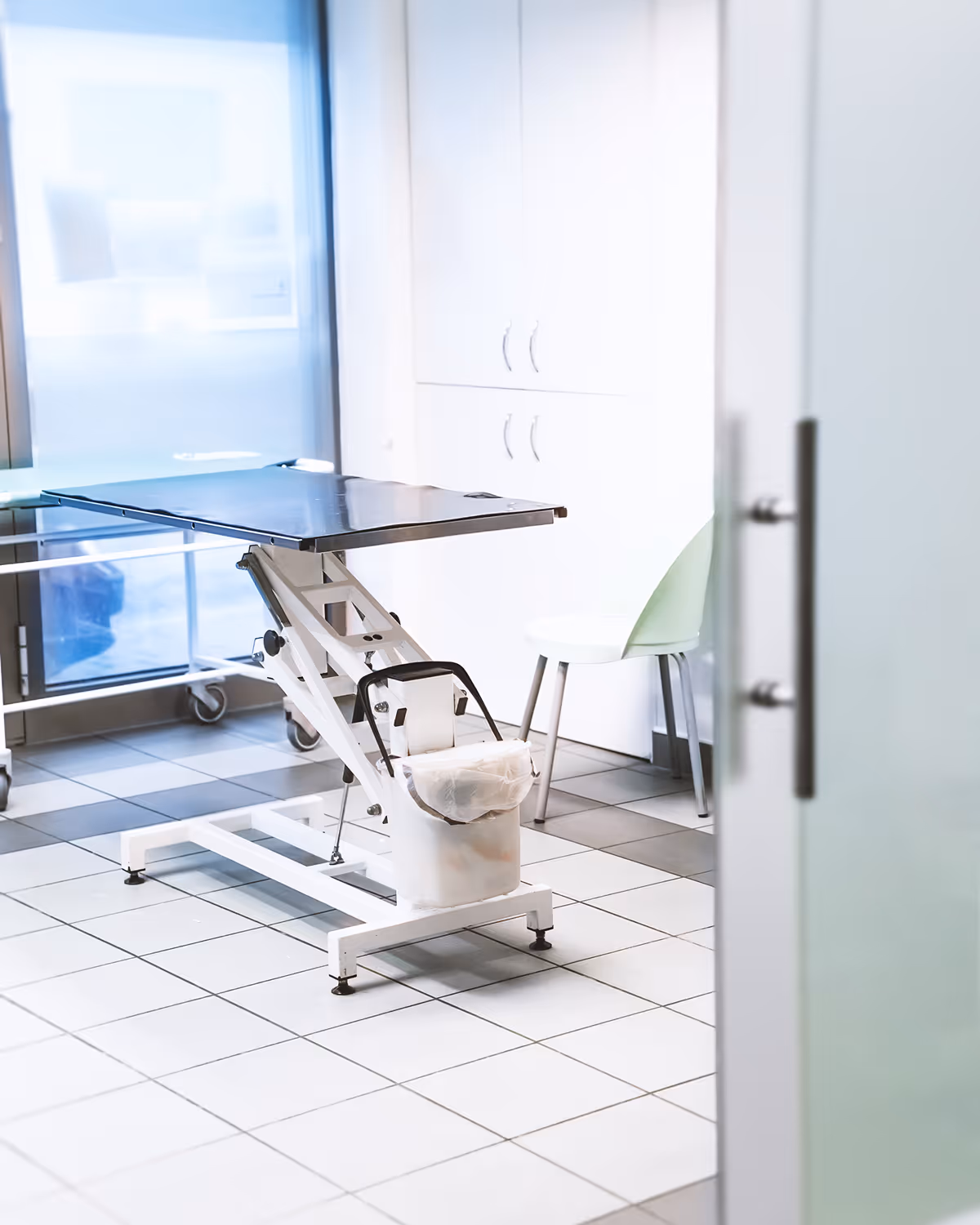Vyšetřovací stůl a židle v čisté klinické místnosti s bílými dlaždicemi a skříňkami.