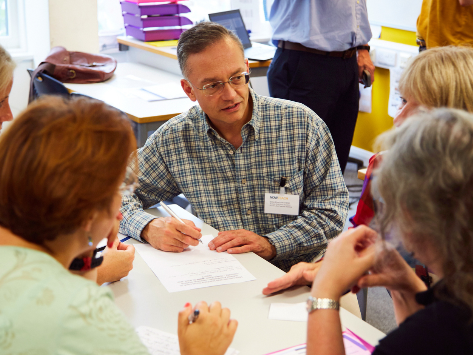 Man writing on a document while sitting at a table with colleagues at a Now Teach event.