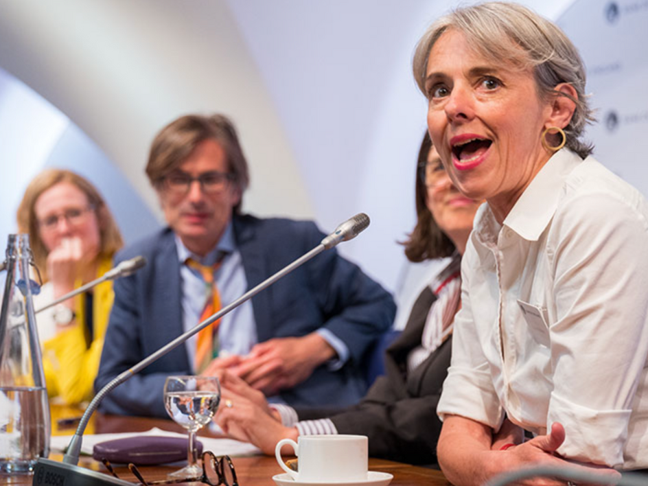 Middle-aged woman speaking animatedly at a panel discussion with people listening behind her.
