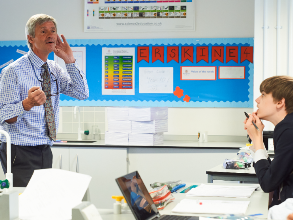 Teacher in a classroom explaining to a student who listens attentively with a pen near his mouth.
