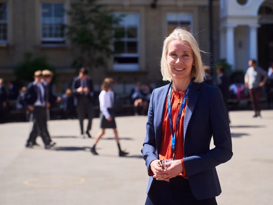 Smiling woman in blazer with staff lanyard stands in schoolyard with students blurred in the background.