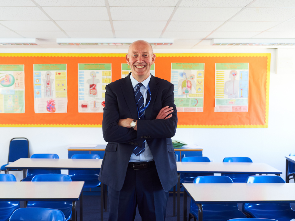 Smiling male teacher in suit standing with arms crossed in an empty classroom with blue chairs.