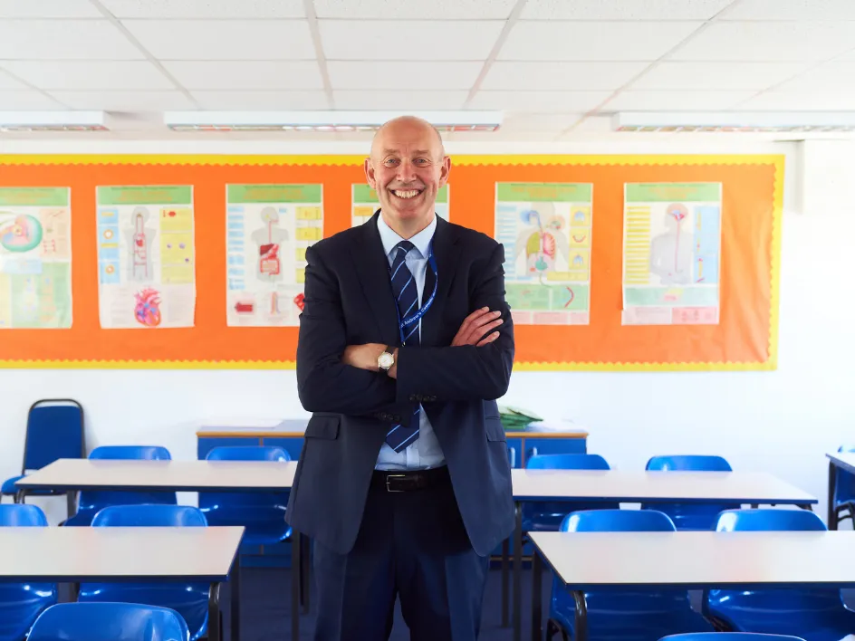 Smiling male teacher in suit standing with arms crossed in an empty classroom with blue chairs.