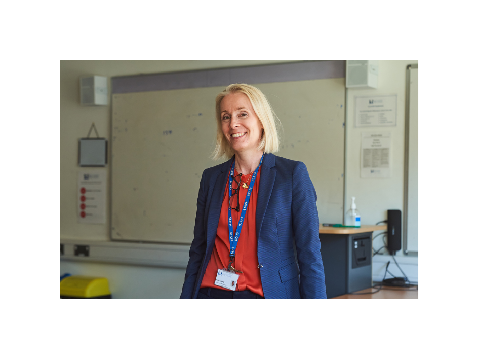 Smiling woman with blonde hair wearing a navy blazer and lanyard, standing in a classroom in front of a whiteboard.