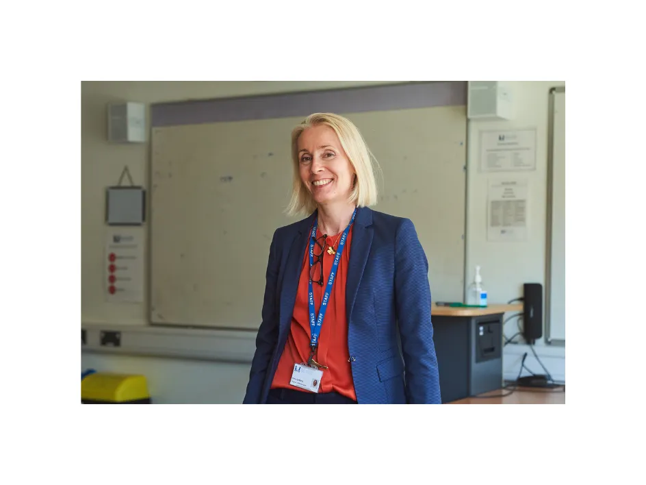 Smiling woman with blonde hair wearing a navy blazer and lanyard, standing in a classroom in front of a whiteboard.