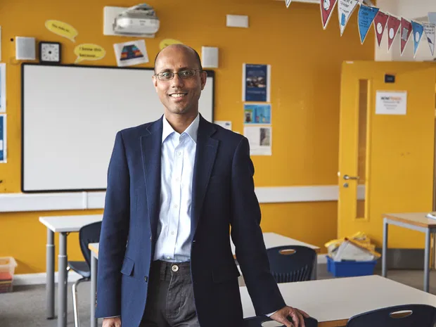 Man in glasses and a navy blazer stands relaxed in front of a bright yellow classroom with desks and bunting.