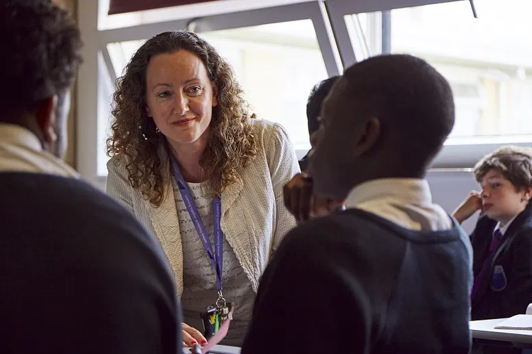 Female teacher with curly hair leans in attentively to speak with secondary school pupils at their desks.