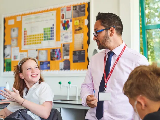 Male teacher with glasses and lanyard engages warmly with a smiling female pupil in a science classroom.