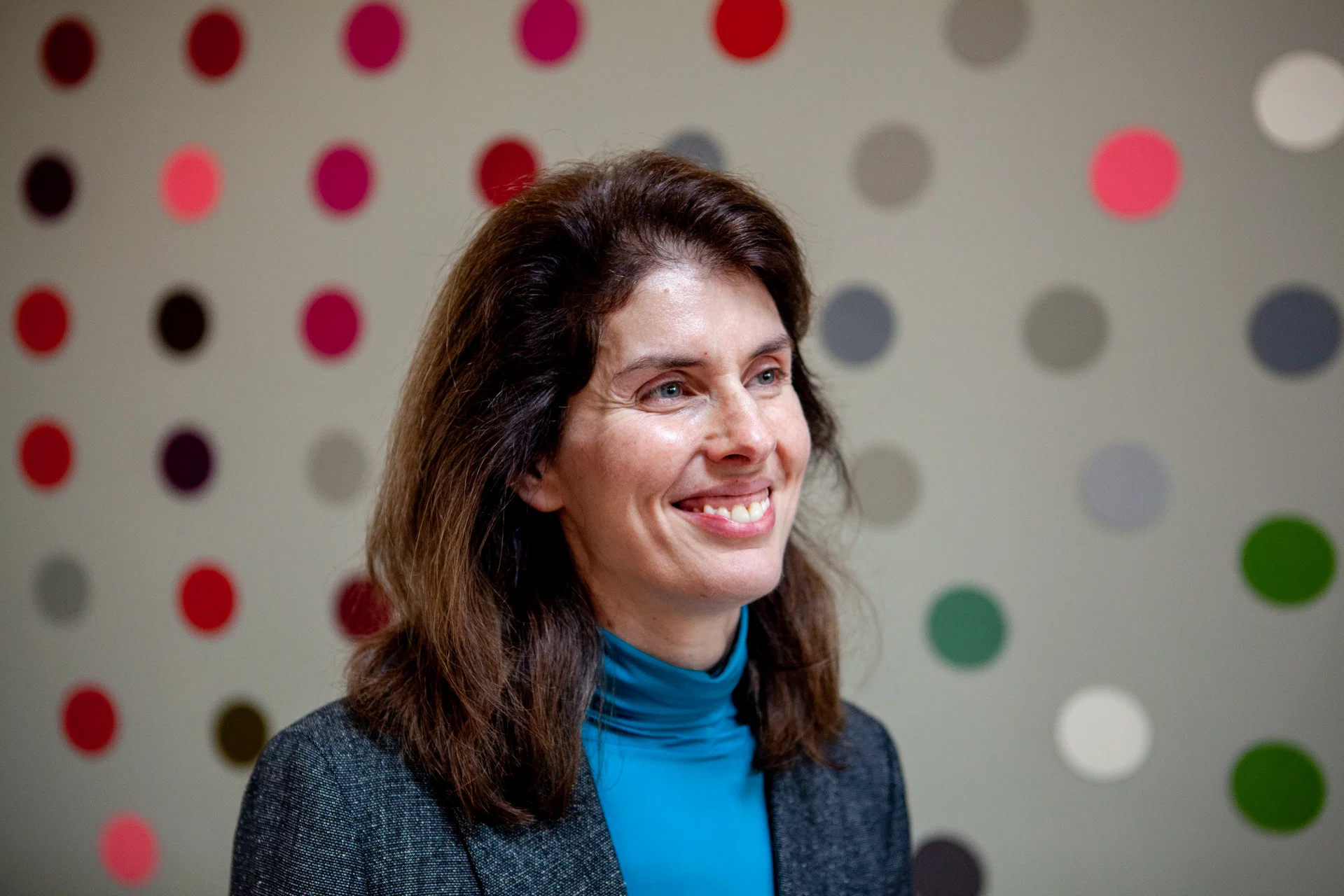Woman with dark hair in a blue turtleneck and grey jacket smiling against a colourful polka-dot background.