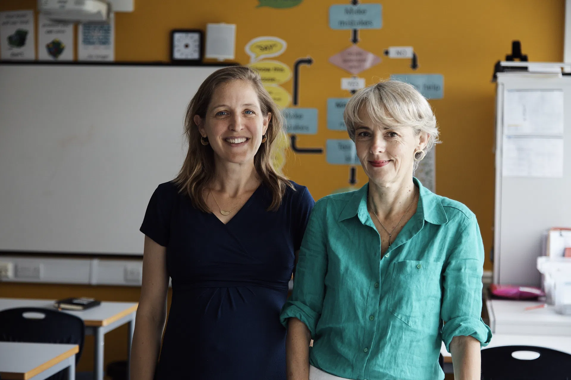 Two women smiling together in a bright classroom with yellow walls and educational displays.
