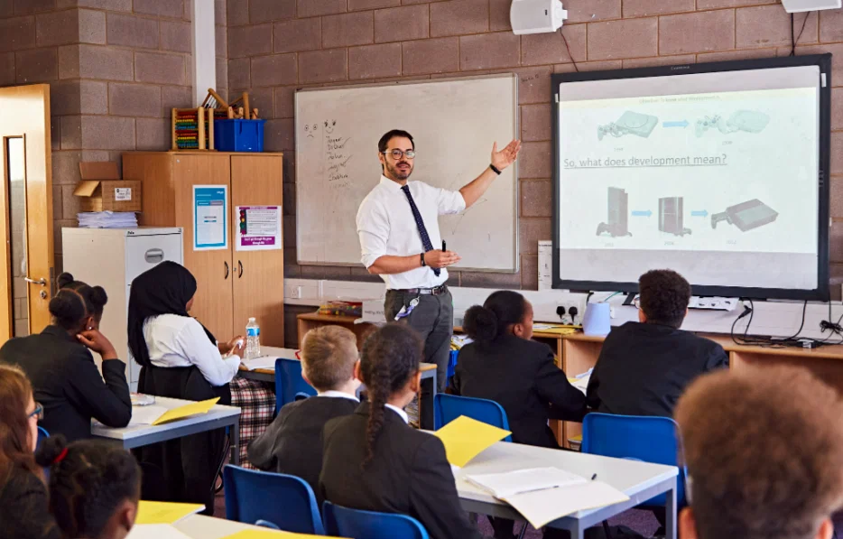 Teacher in white shirt and tie instructs diverse students seated in desks facing a projector screen.