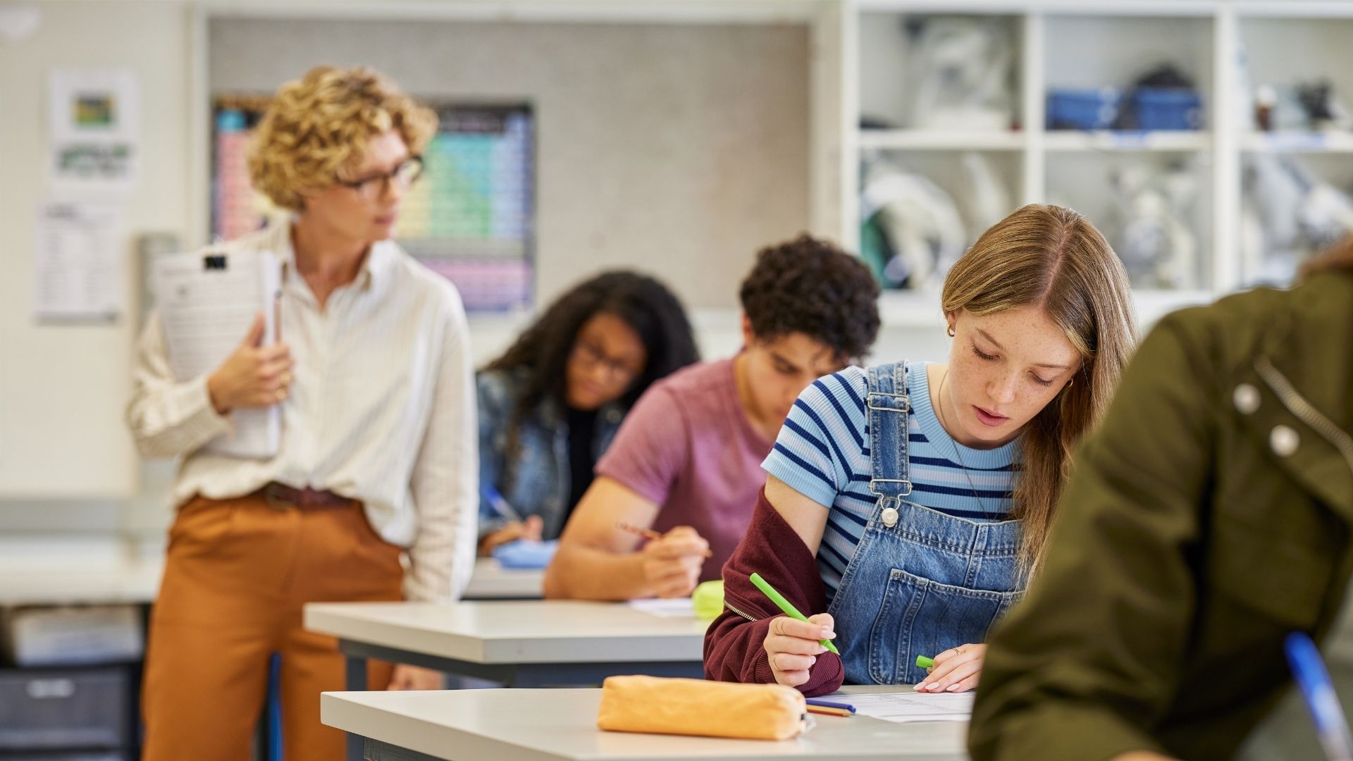 Lehrerin geht durch das Klassenzimmer, während Schülerinnen und Schüler schreiben
