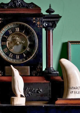 Black vintage clock with Roman numerals, a small ceramic penguin, a red and black decorative column, and a curved white object labeled 'ANTARCTICA 19 37.32' on a shelf against a green wall.