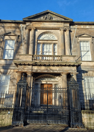 Historic stone building with classical columns, a gated black iron fence, and arched windows under a clear blue sky.