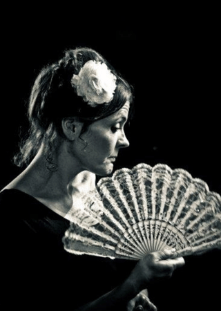 Profile of a woman with a white flower in her hair holding a lace fan against a black background.