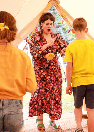 Woman in a floral dress and green sandals interacting animatedly with two children standing inside a tent.