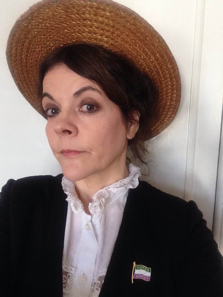 Woman wearing a brown straw hat, white ruffled blouse, and black jacket with a 'Votes for Women' suffragette pin.