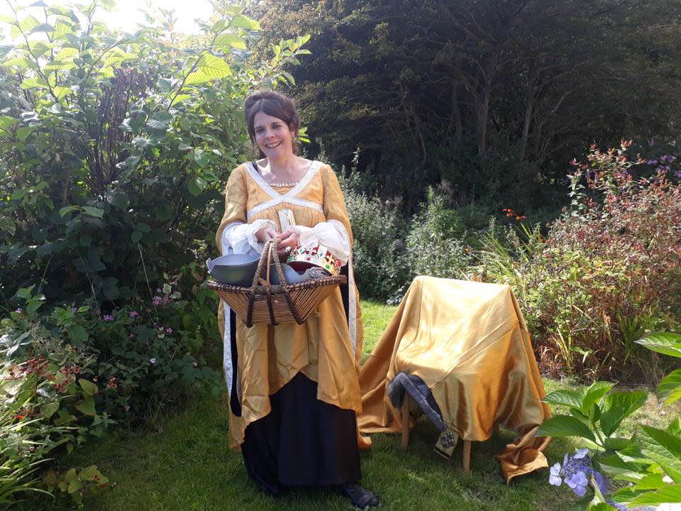 Woman in a yellow historical dress holding a basket with fabric and a crown, standing outdoors on grass near a chair covered with golden cloth.