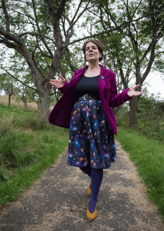Woman in a purple blazer and galaxy-patterned skirt standing on a grassy path with trees in the background.