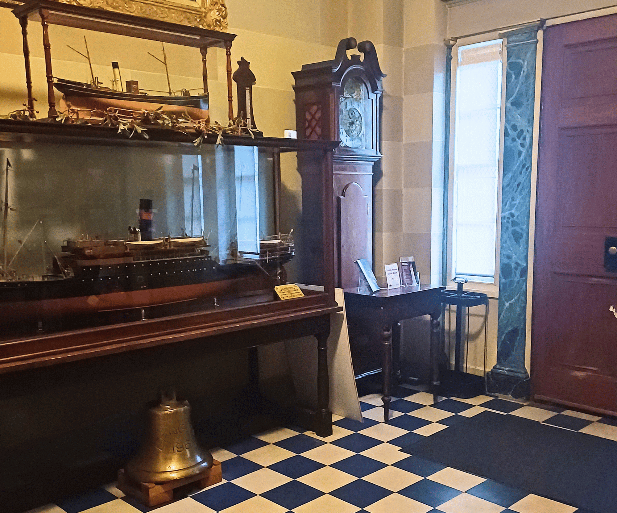 Room with ship models in glass case, grandfather clock, small table, bell on checkerboard floor.