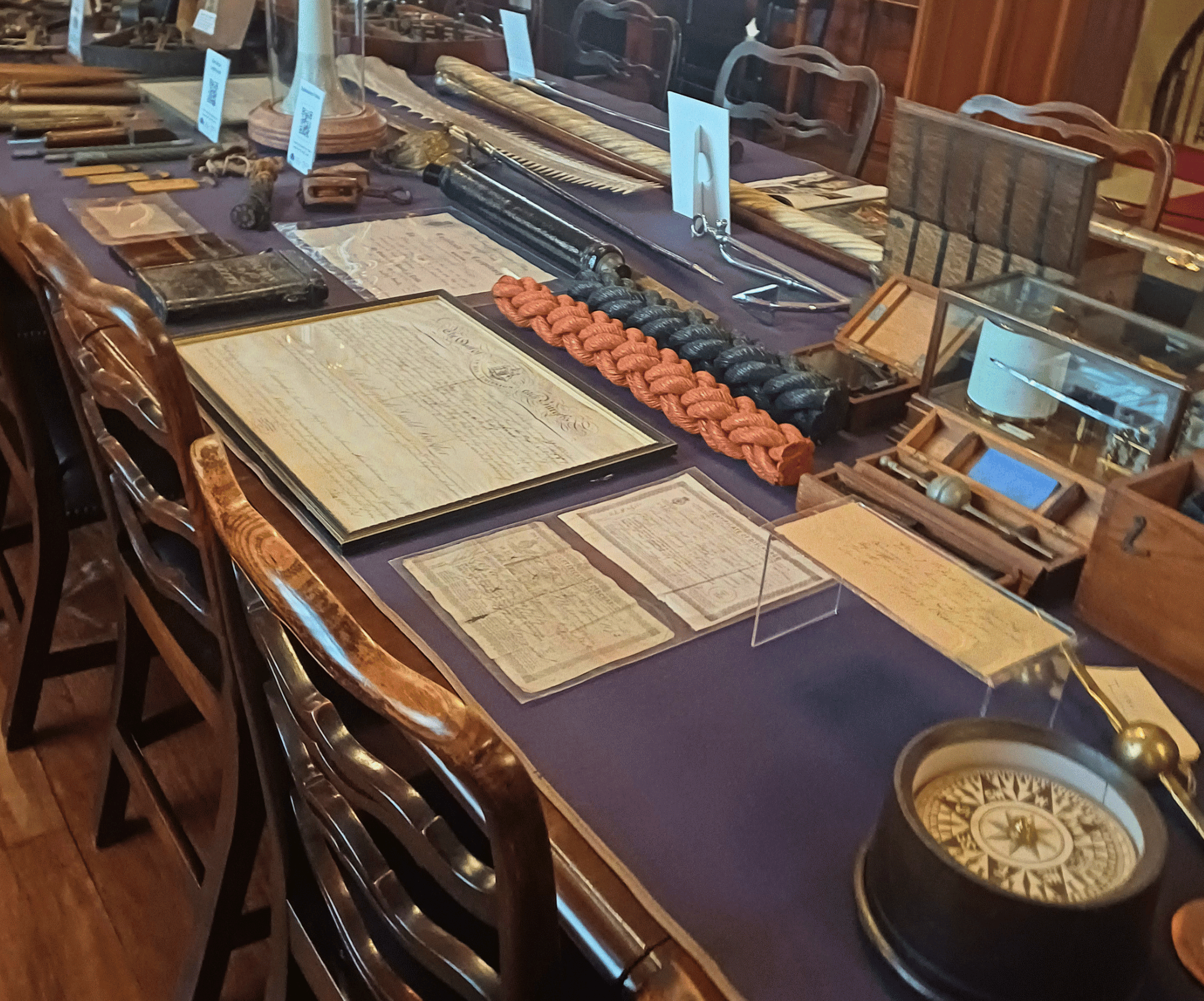 Museum display of antique documents, ropes, nautical tools, and a compass on a wooden table with chairs.