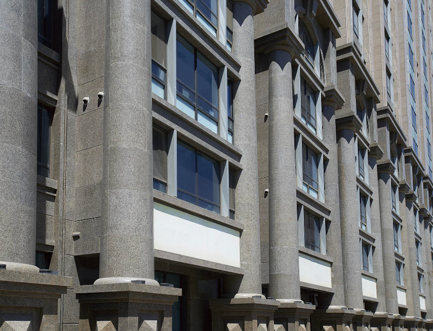 A close up shot of a grey stone building exterior featuring large cylindrical columns alongside a grid of rectangular windows with dark grey frames.