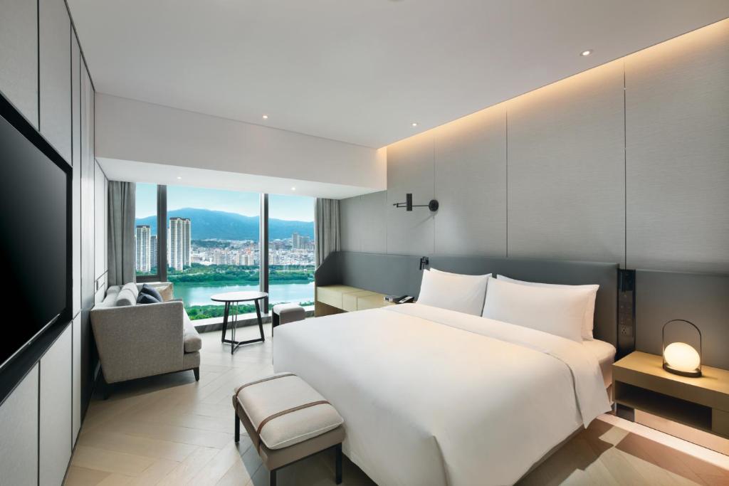 A minimalist hotel room featuring a white king-size bed, a grey panelled headboard wall with a black swing-arm lamp, and a floor-to-ceiling window offering views of a river and city skyline.