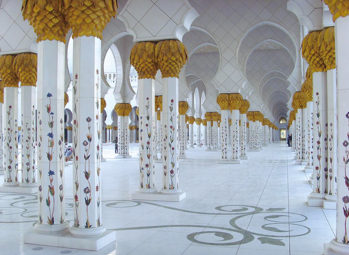 A grand hotel lobby featuring a polished marble floor with a large dark brown geometric inlay pattern leading towards a reception desk and large pillars.