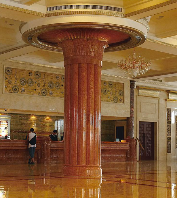 An ornate hotel lobby featuring a large fluted marble column in a reddish orange hue with a decorative papyrus style capital that meets a circular recessed ceiling.