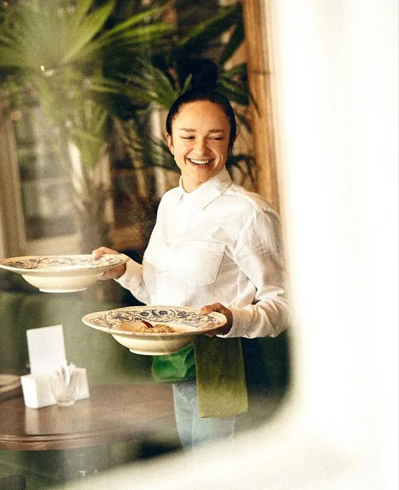 Smiling waitress carrying two decorative plates with food in a restaurant setting.