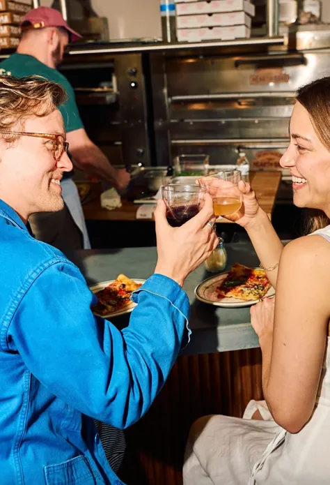Two people clinking glasses and eating pizza at a counter with a cook in the background.