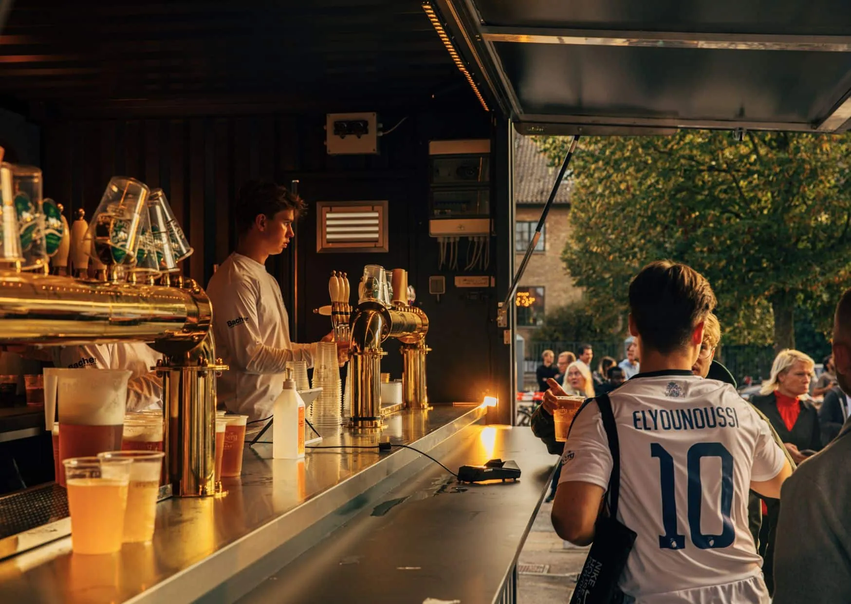 Barista serving drinks to people at an outdoor beer stand with golden taps during sunset.