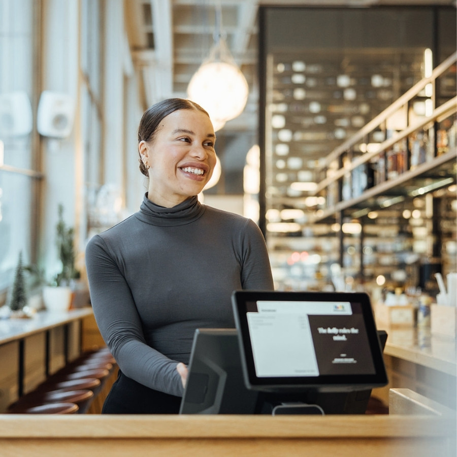 Smiling woman stands behind a restaurant POS system counter in a modern cafe.