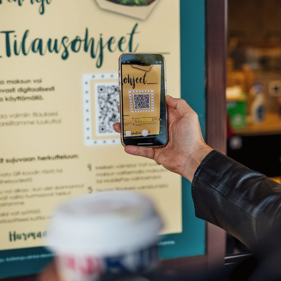 Person scanning a QR code on a yellow sign using a smartphone, holding a coffee cup.